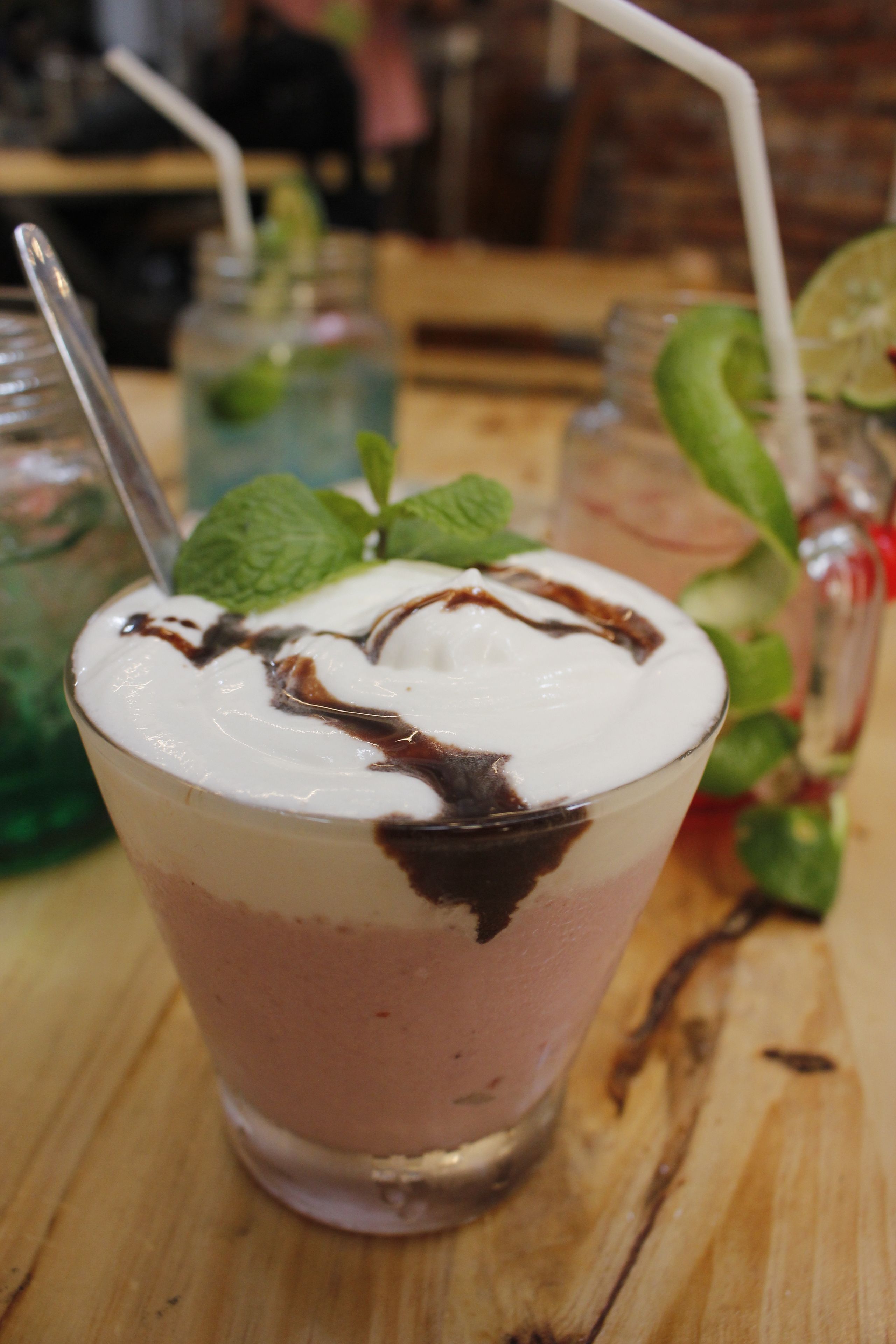 Een glas gelaagde drank met opgeklopt room en chocoladesiroop, versierd met muntblaadjes, op een houten tafel.