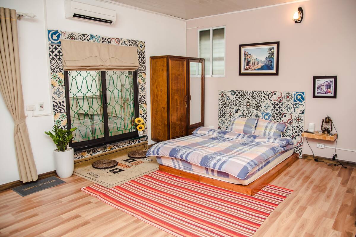 A bedroom with a bed, a window with curtains, a wardrobe, a bedside table, and a wall-mounted air conditioner.