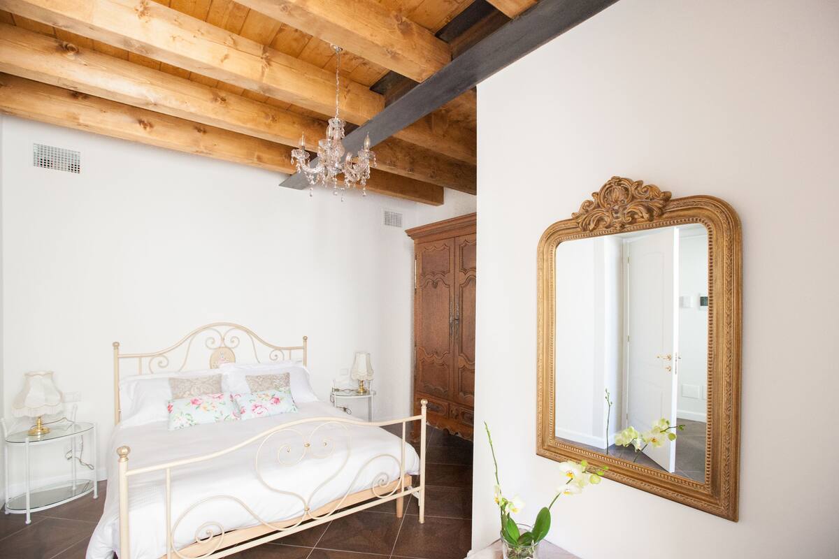 A bedroom with a wooden ceiling, a white bed, a wooden wardrobe, and a large mirror on the wall.