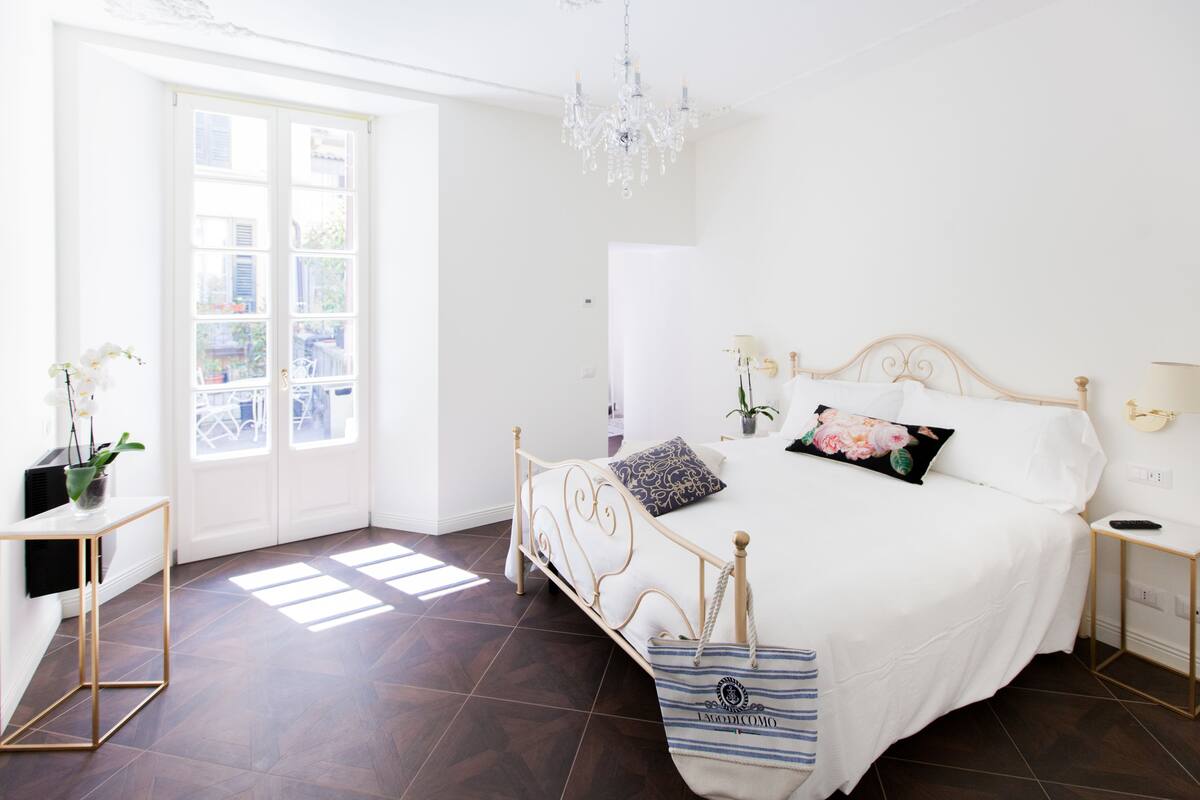 A bedroom with a bed, bedside tables, a chandelier, a window, and a door.