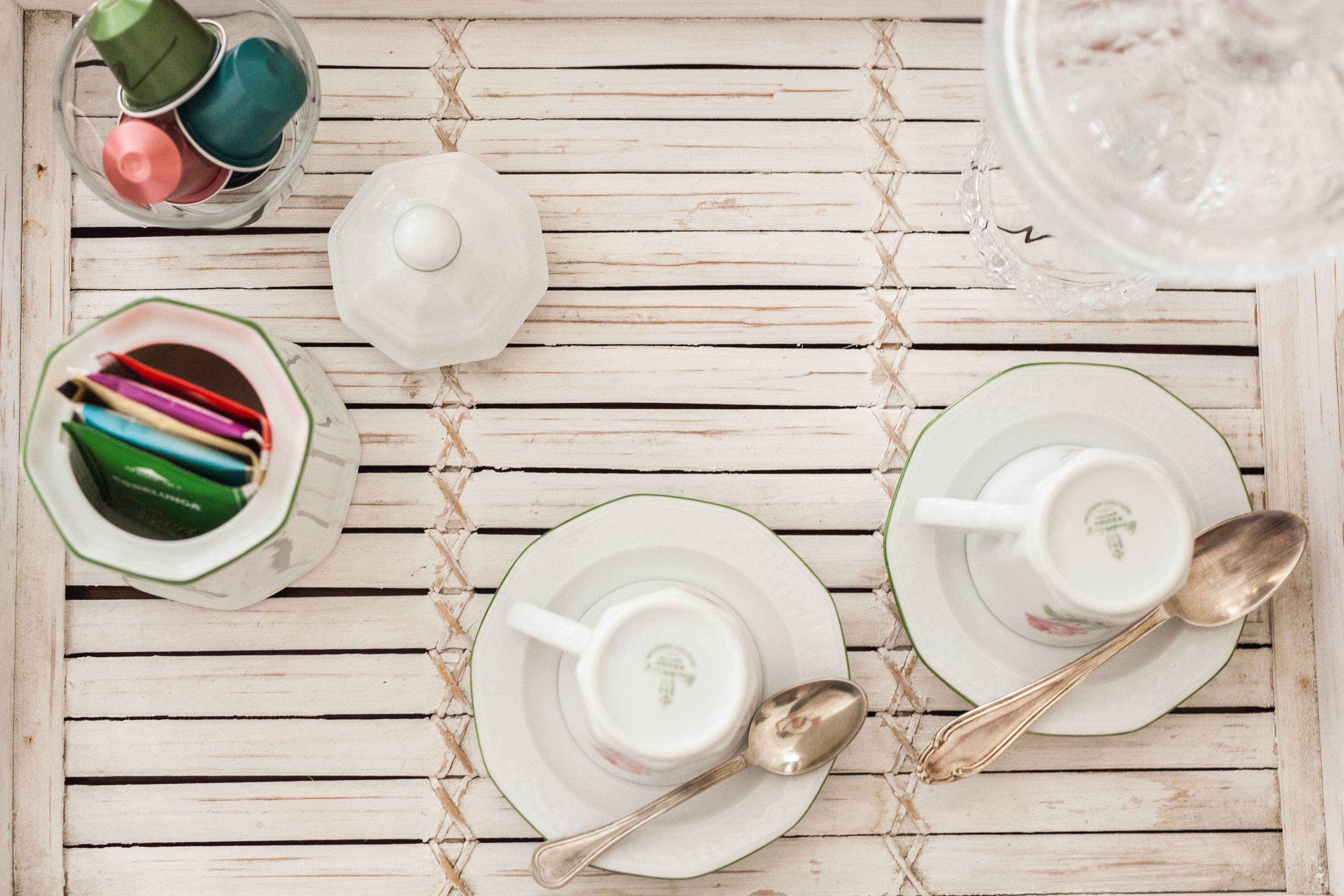 Una mesa de madera blanca con un hervidor blanco, dos tazas blancas con sus platitos, dos cucharas y un tarro de cristal.