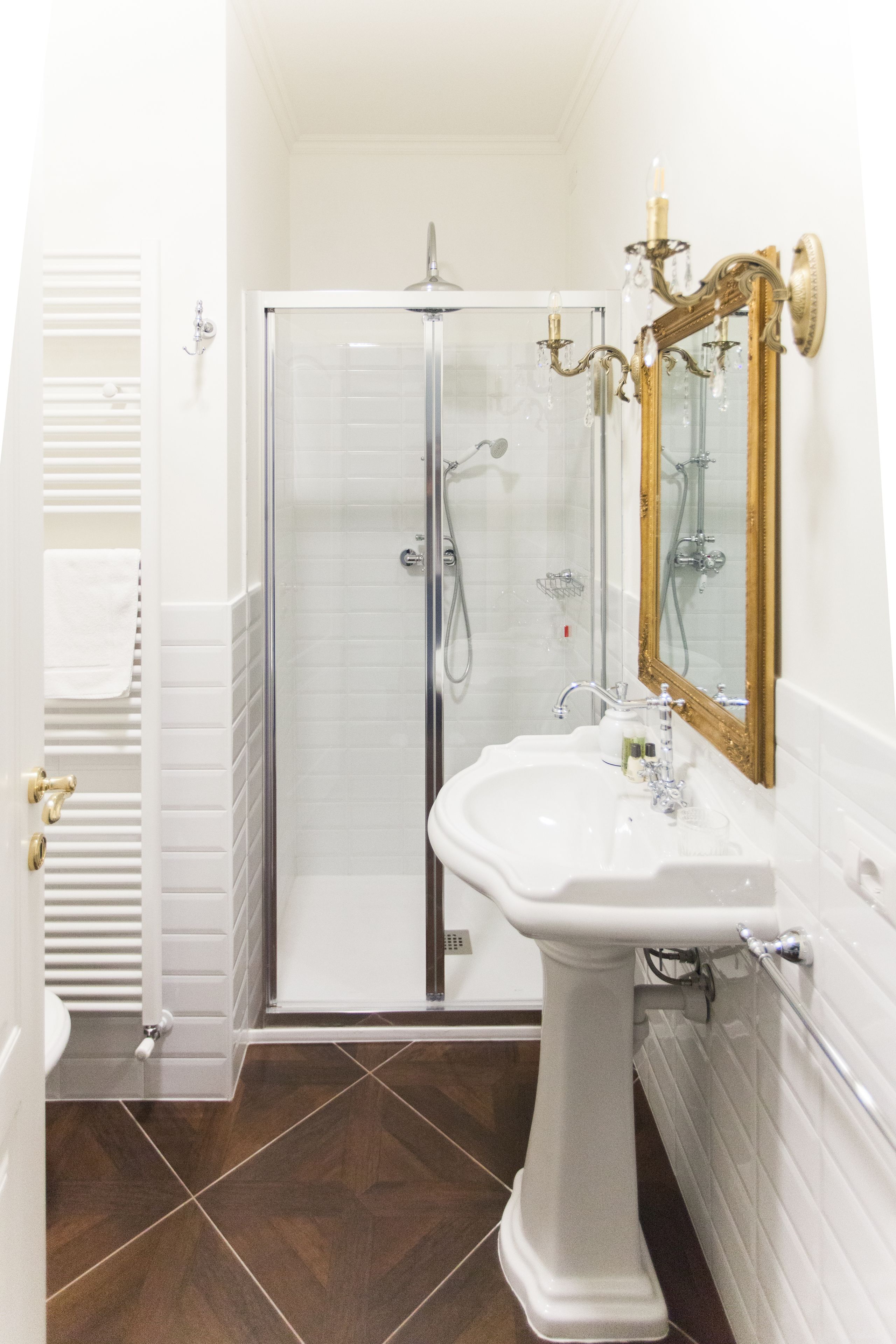 Un baño con cabina de ducha de vidrio, lavamanos pedestal y un espejo grande con marco dorado.