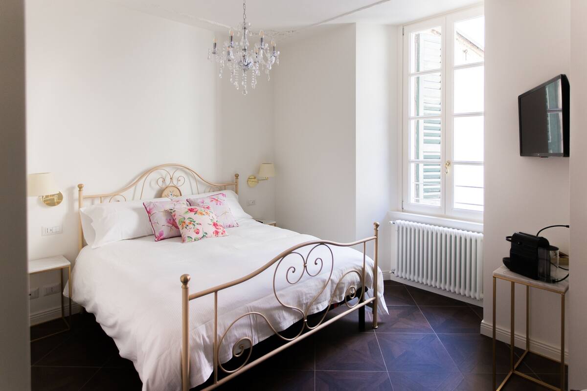 A bedroom with a bed, a chandelier, a window, a radiator, and a television on a stand.