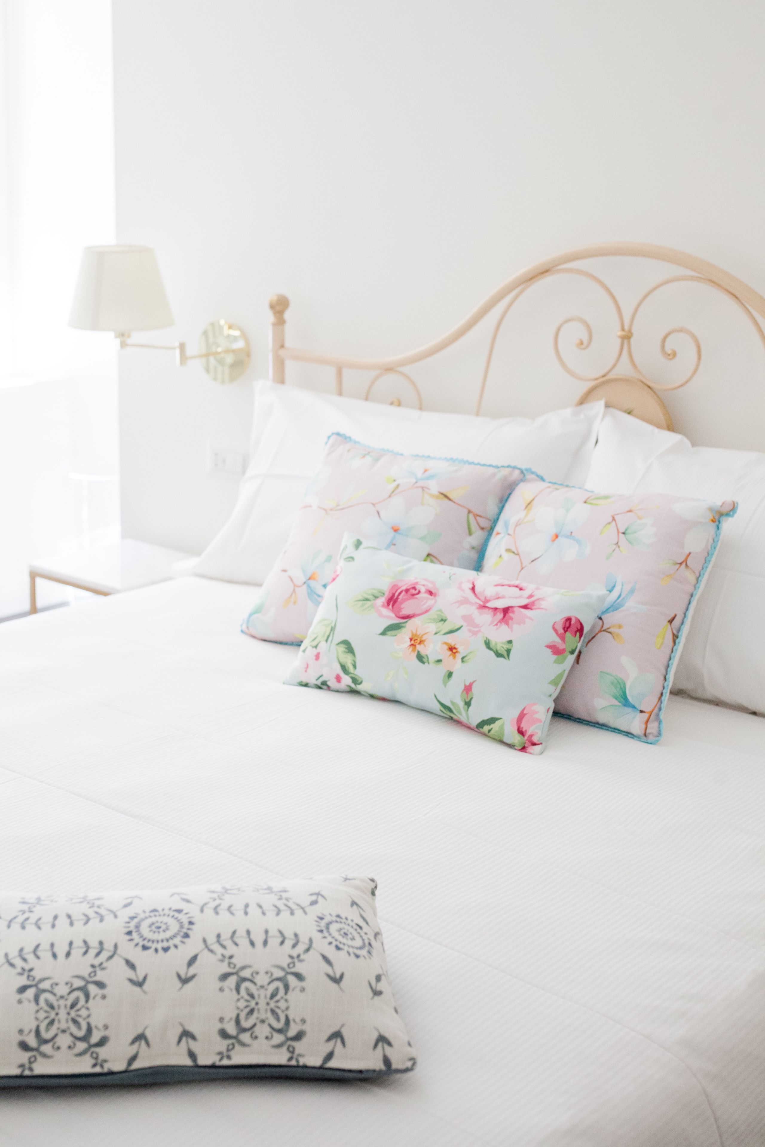 Una cama bien hecha con almohadas estampadas y con motivos florales, una mesita de noche con lámpara, y un aplique en la pared.