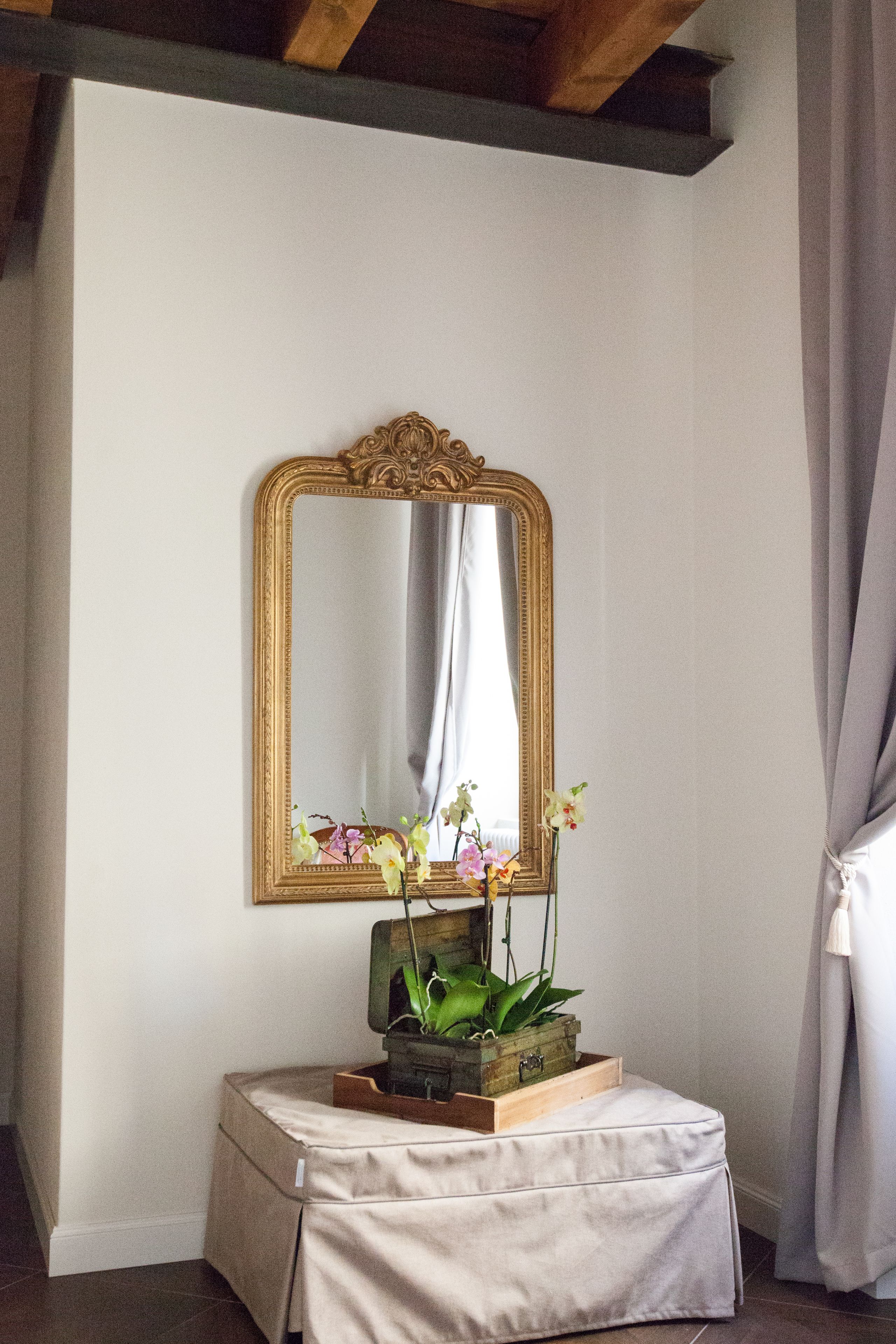 Une pièce avec un grand miroir décoratif au mur, une petite table sur laquelle est posée une plante, et un rideau à la fenêtre.