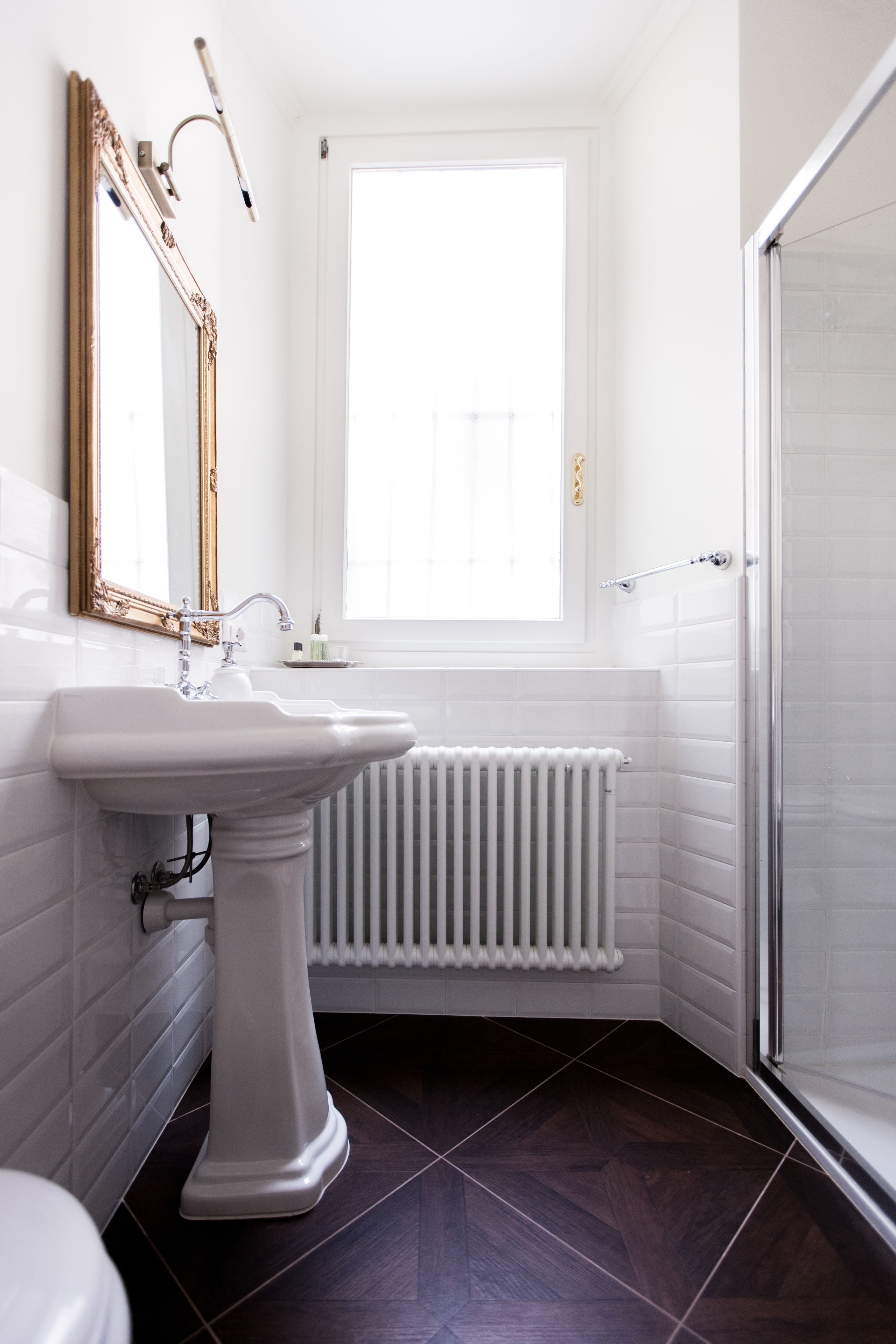 A bathroom with a pedestal sink, a large mirror, a window, a radiator, and a shower area.