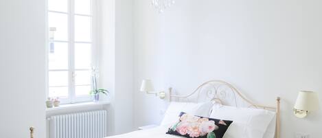 Un dormitorio con una cama blanca, un candelabro, una ventana con vistas y un radiador.