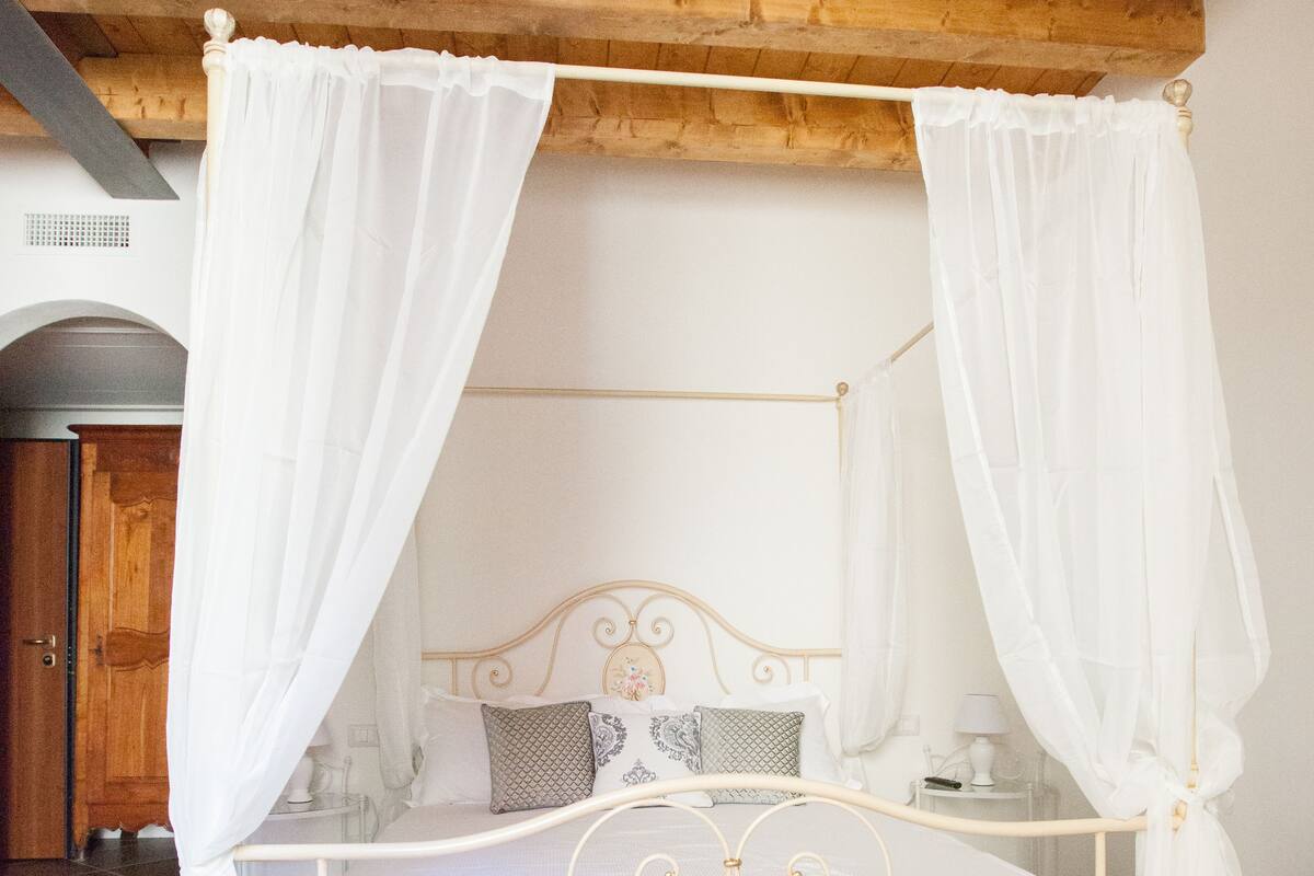 A four-poster canopy bed with a metal frame and white bedding, set in a room with wooden ceiling beams and dark tiled flooring.