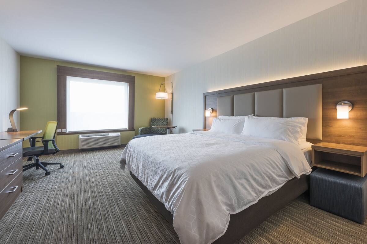 A hotel room with a large bed, a desk with a chair, a window, and a wall-mounted light fixture.