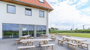 Une terrasse extérieure dotée de plusieurs tables de pique-nique en bois et d’un bâtiment avec de grandes fenêtres et un toit rouge.