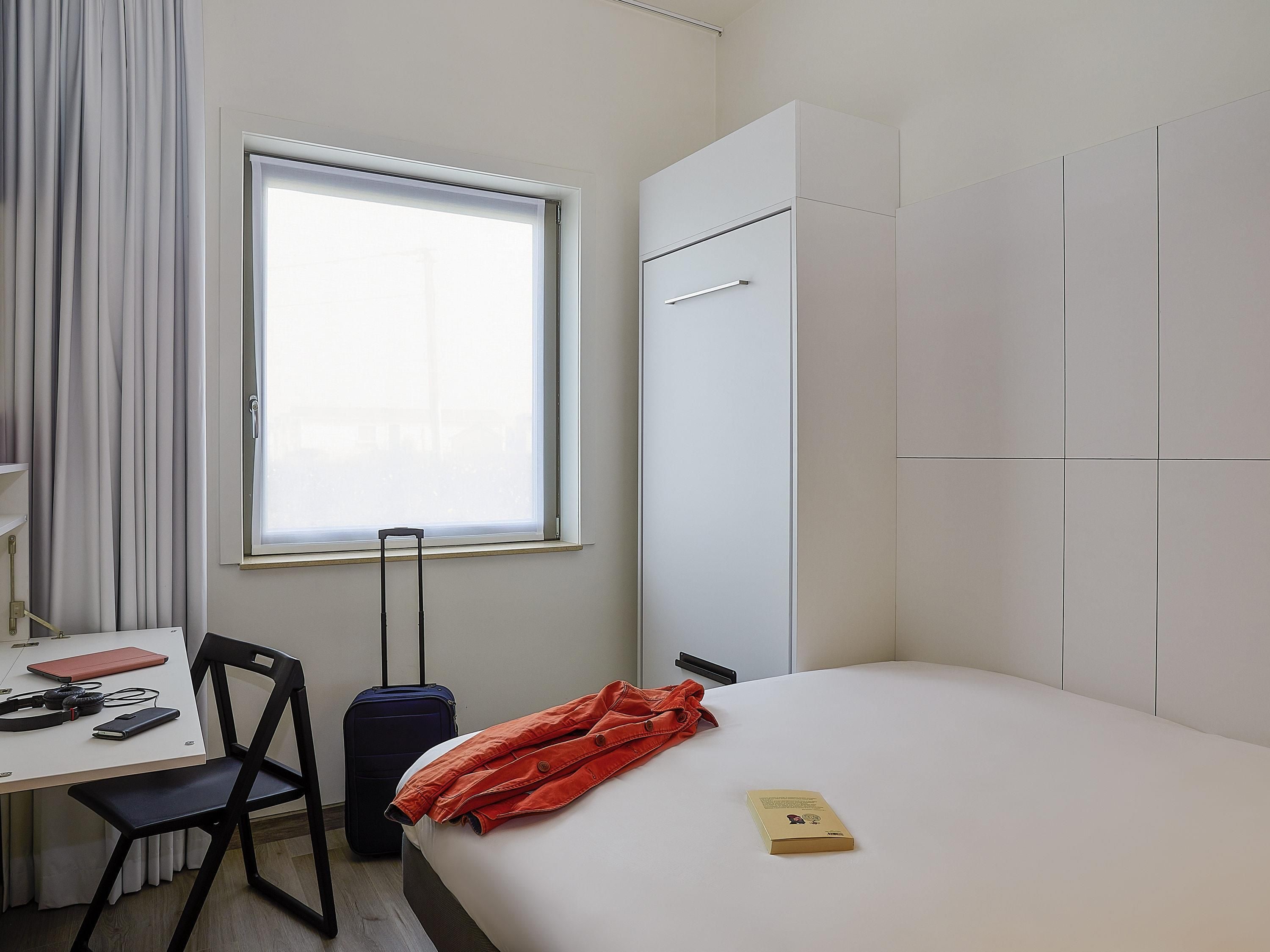 Quarto de hotel com cama, escrivaninha, cadeira, mala e janela.