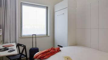 Quarto de hotel com cama, escrivaninha, cadeira, mala e janela.