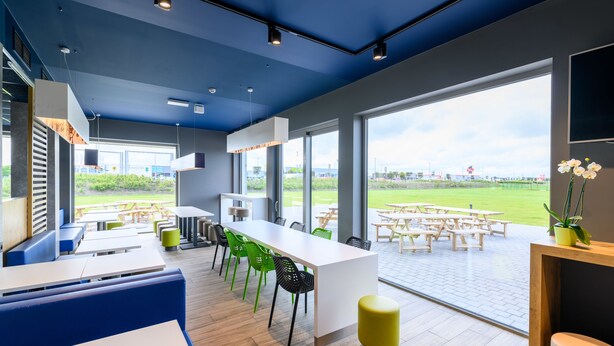 Un espace intérieur moderne avec une longue table blanche, des chaises vertes et un tabouret jaune. De grandes fenêtres offrent une vue sur un espace extérieur aménagé avec des tables et des chaises.