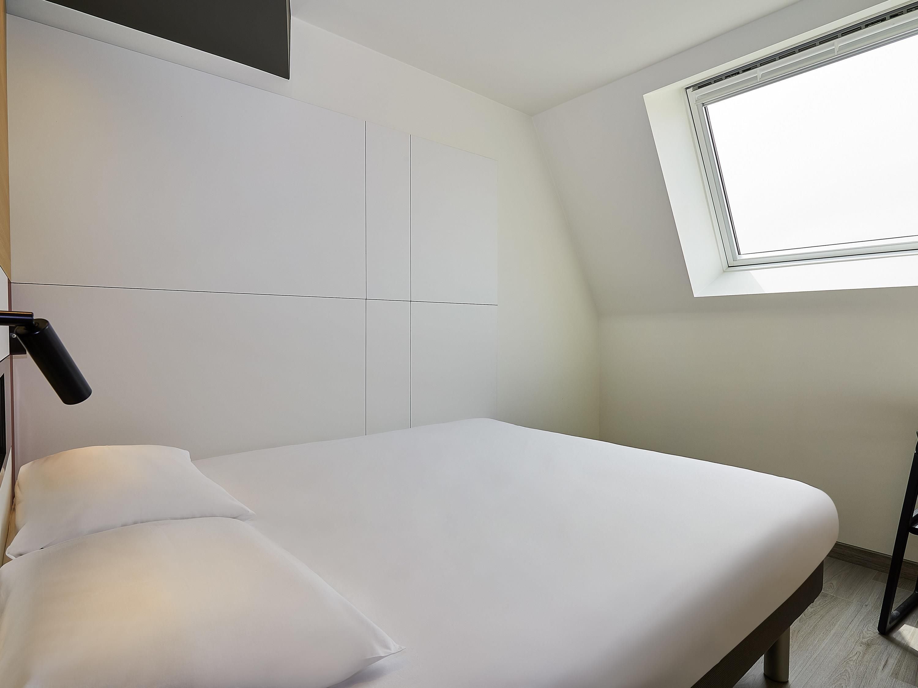 Un dormitorio con cama, lámpara de noche, ventana y televisor en la pared.