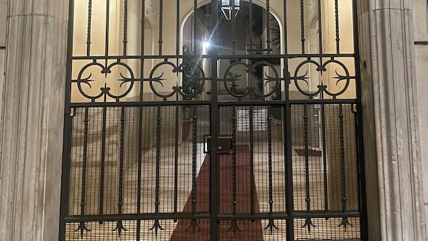 Una reja de hierro forjado con diseños intrincados, una placa con un nombre en la parte superior y el reflejo de un interior de edificio.