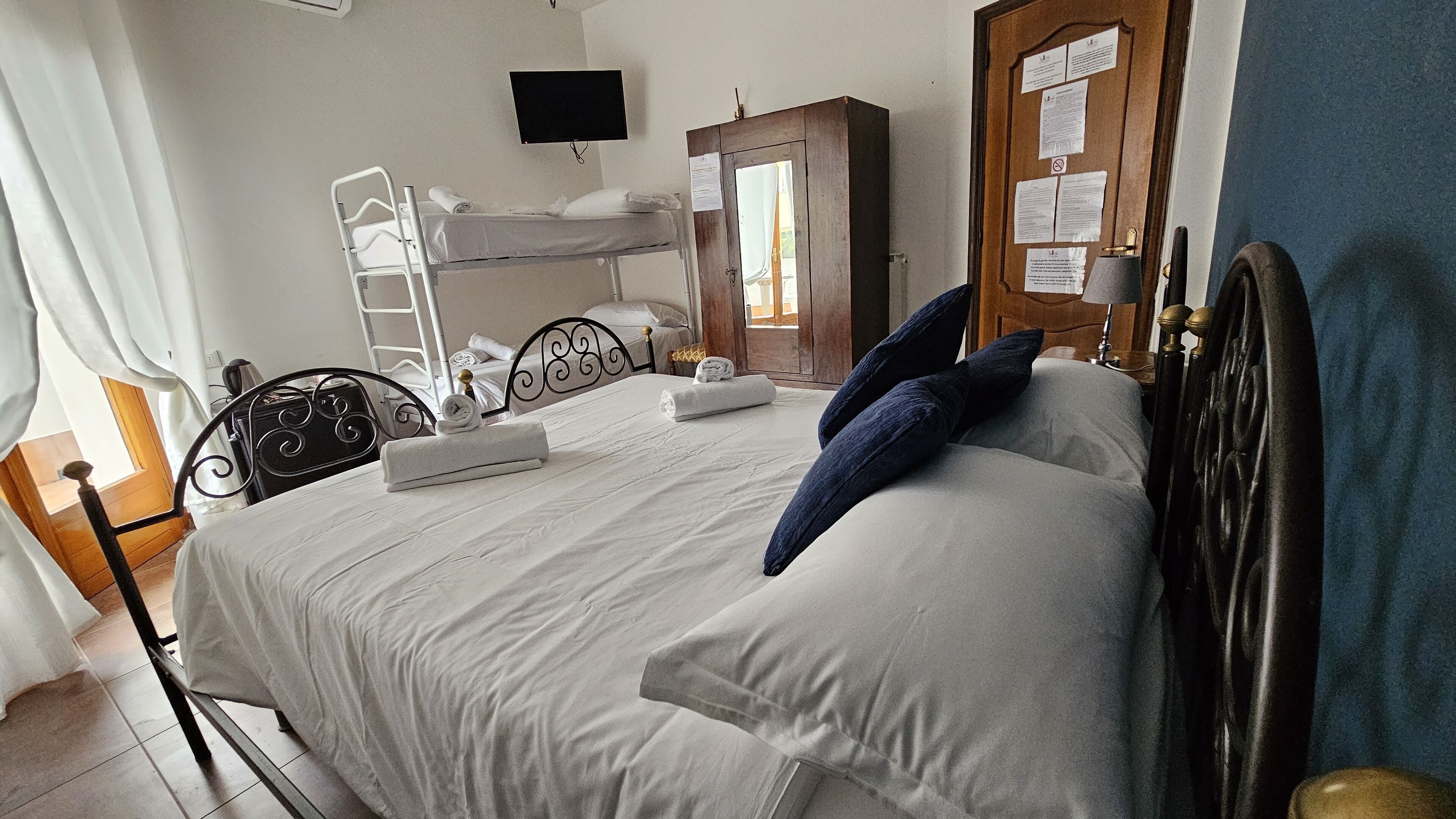 A single bed with a white mattress and blue pillows, a bedside table with a lamp, a wooden wardrobe, and a window with curtains.