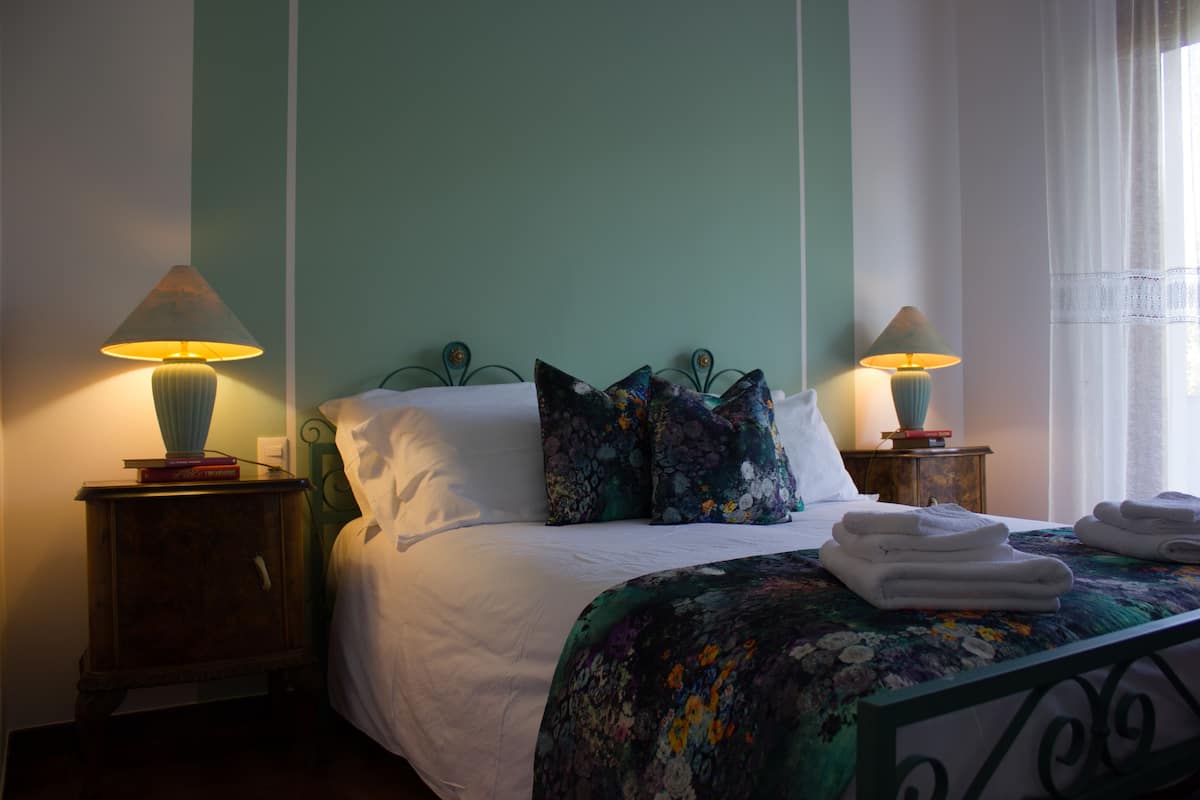 A neatly made bed with a floral bedspread, two bedside lamps, and a small wooden nightstand with books.