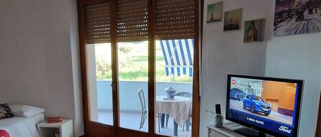 Una stanza con un letto, una TV su una cassettiera, un balcone con tavolo e sedie, e vista sul verde.