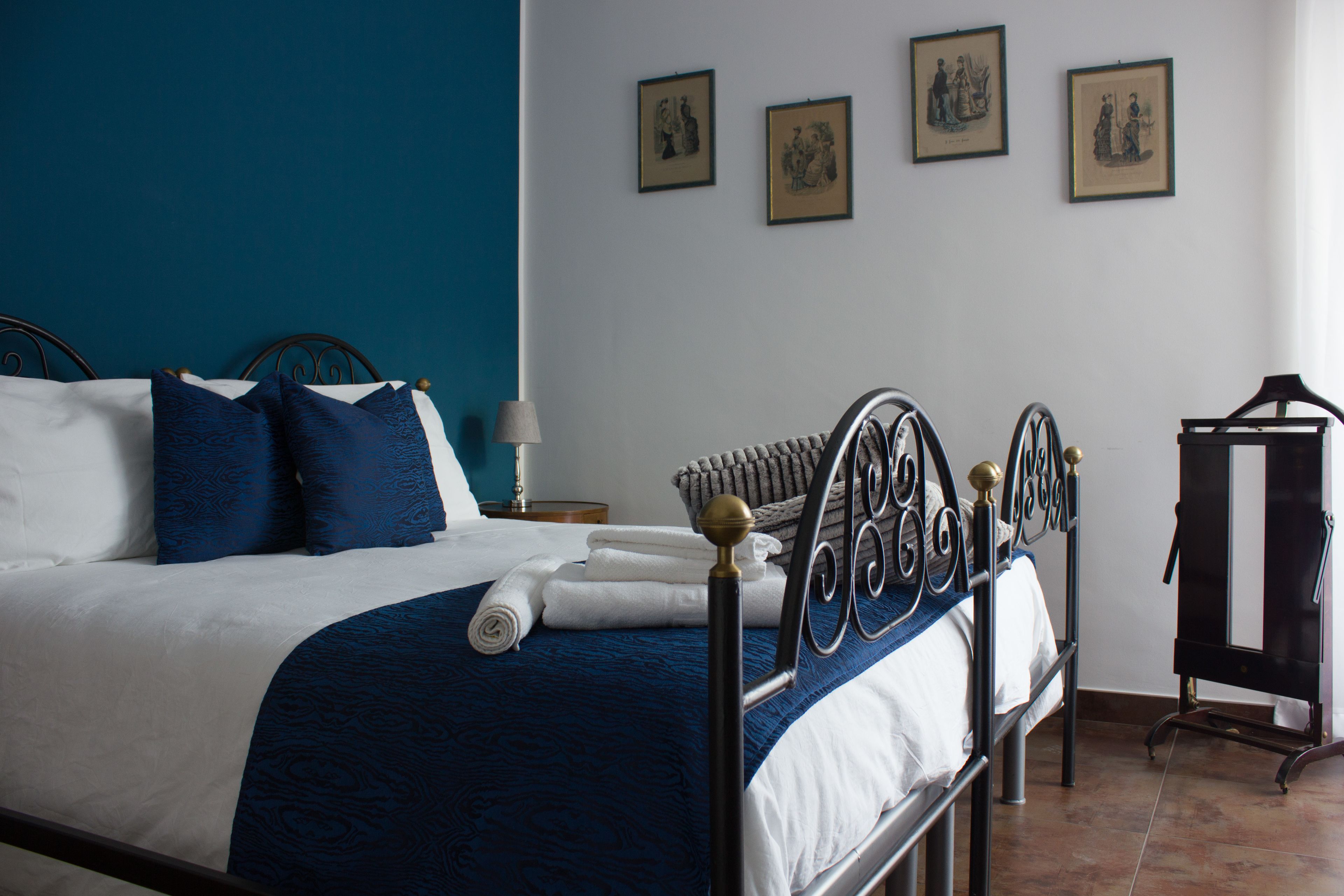Uma cama bem arrumada com colcha azul e branca, cabeceira de metal e mesa de cabeceira com abajur.