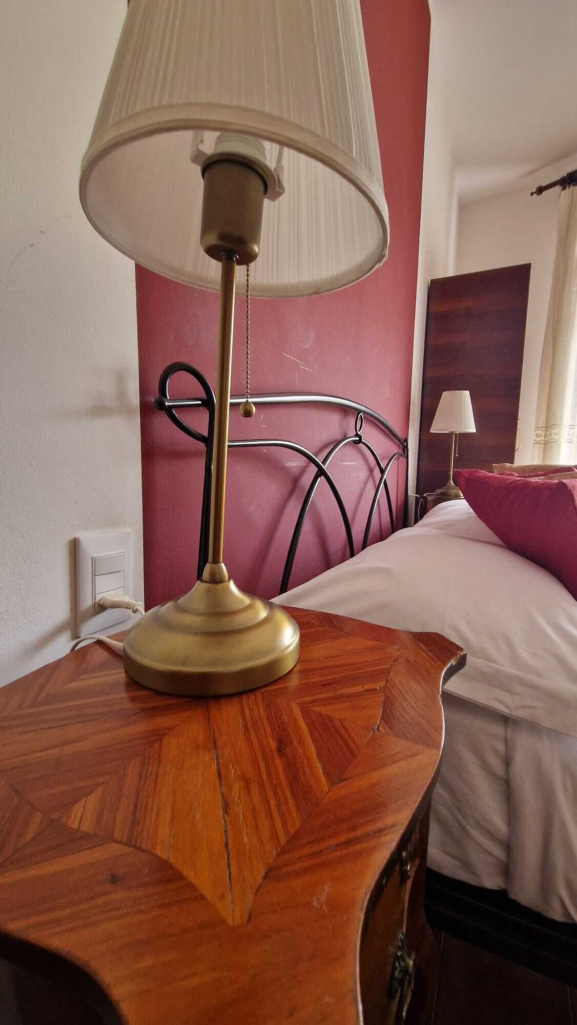 Une table de chevet en bois avec une lampe, un lit avec une tête de lit et une prise électrique murale.
