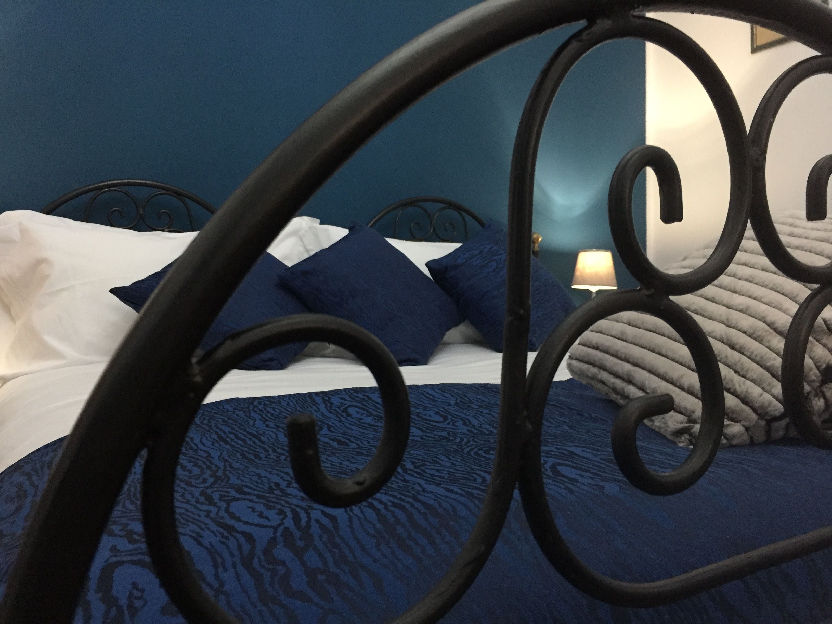 A bed with a blue patterned bedspread and white pillows, a black metal headboard, and a bedside lamp.
