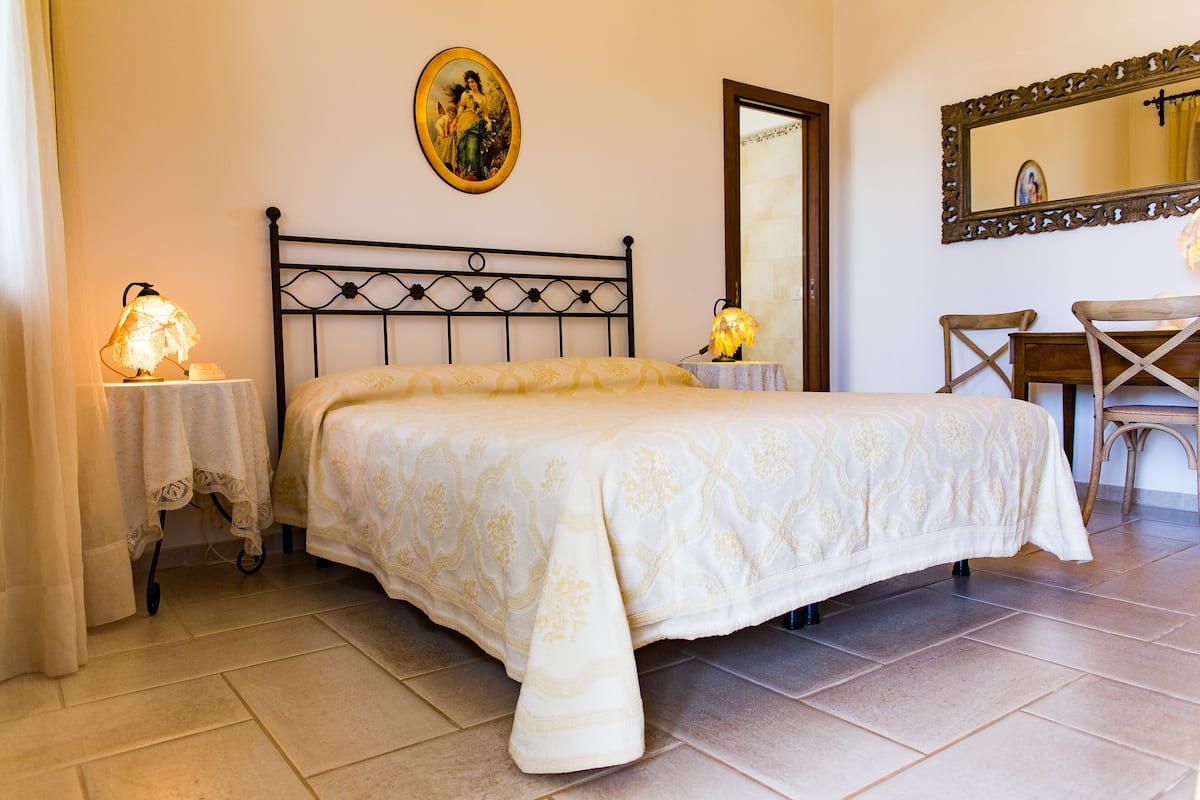 A bedroom with a bed, a nightstand, a table with a lamp, a chair, a wooden desk, a mirror, and a framed picture on the wall.
