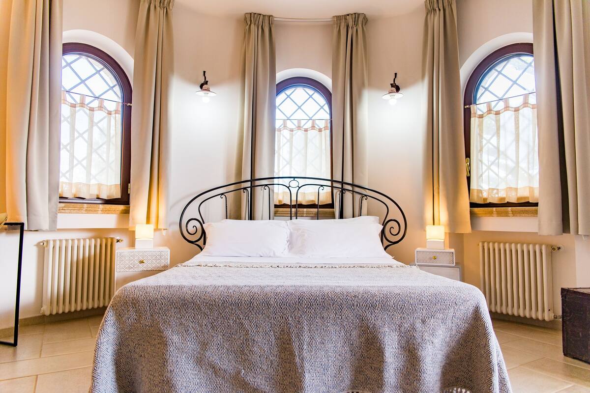A neatly arranged bedroom with a bed centered in the room, flanked by two bedside tables with lamps. There are three arched windows with curtains, allowing natural light to enter.