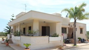 Una casa moderna de dos pisos con un porche cubierto, plantas en macetas y una palmera.