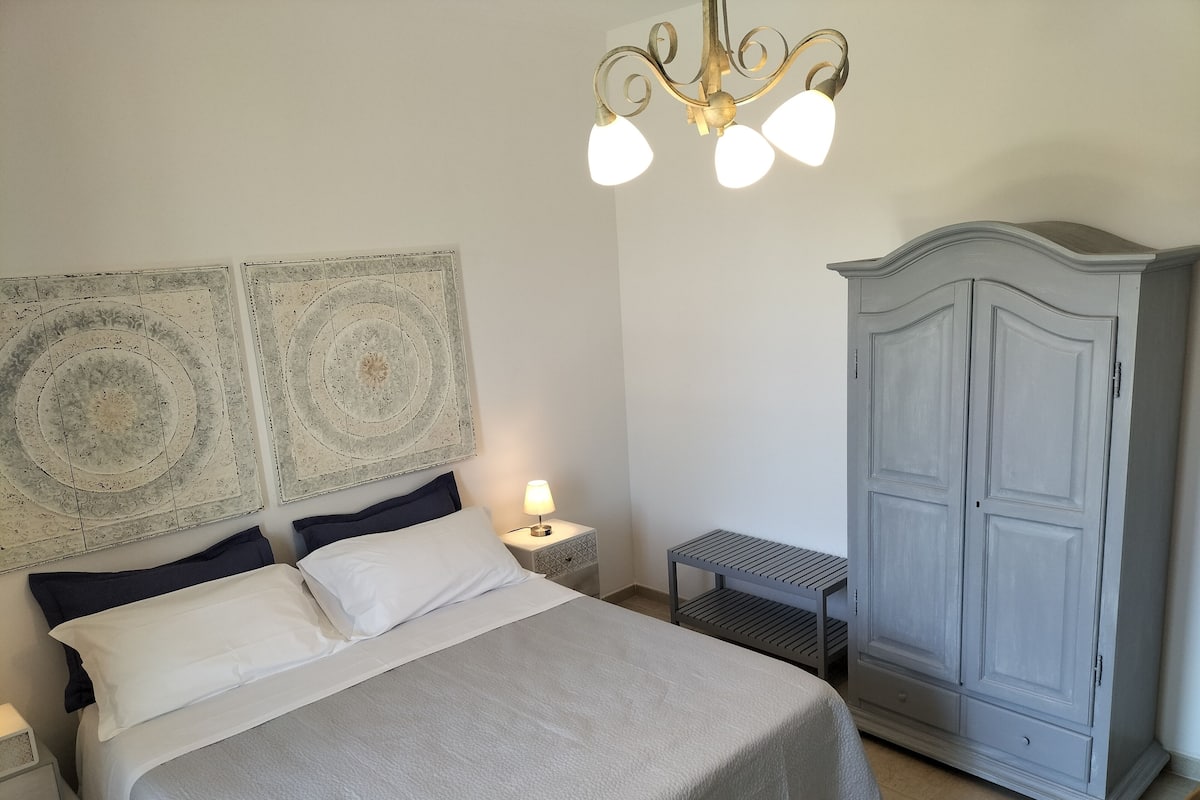 A neatly made bed with a grey bedspread and two pillows, a bedside lamp, a small table, and a wardrobe with a light grey finish.