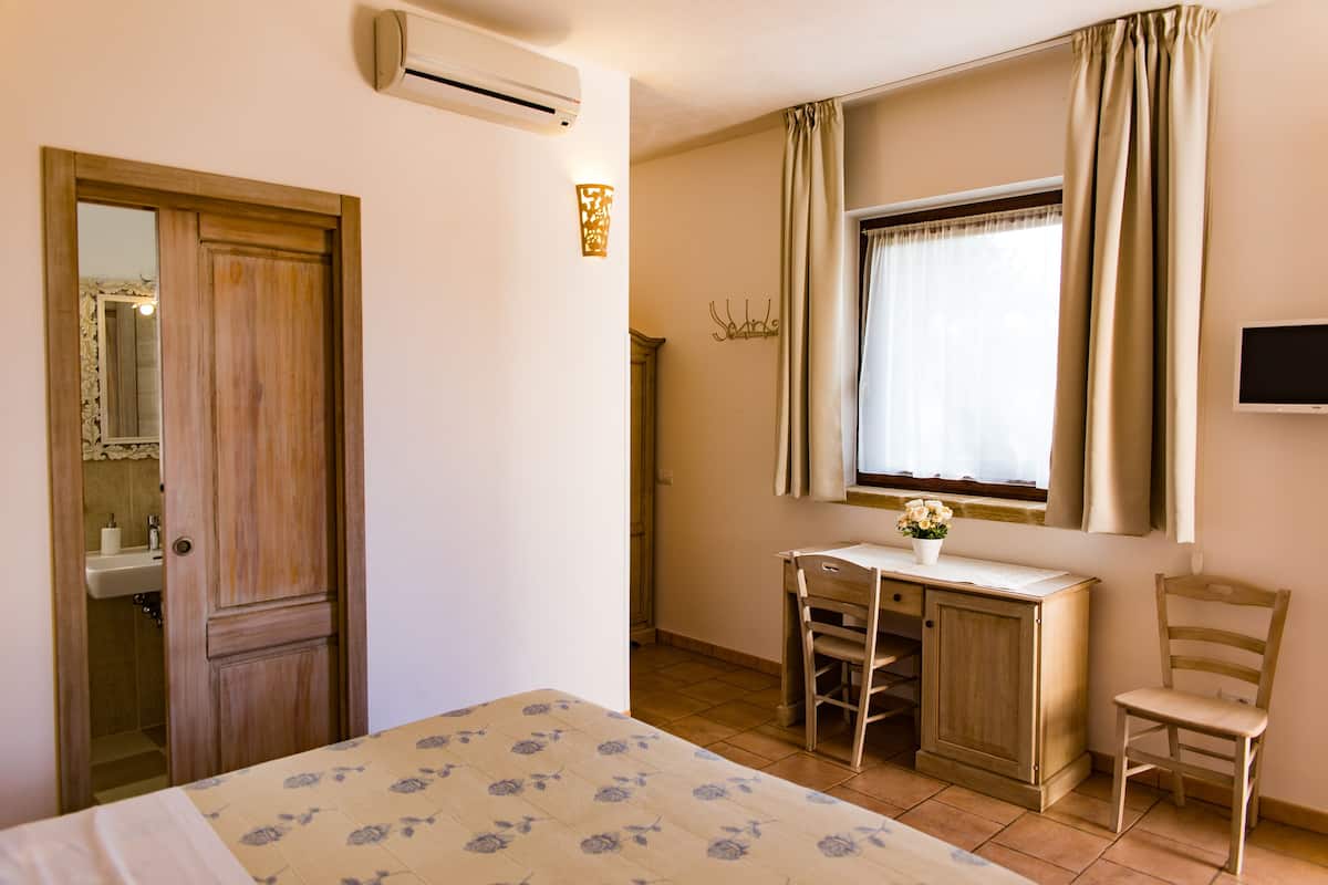 A bedroom with a bed, a desk, a chair, a window with curtains, a mirror, and an air conditioning unit.