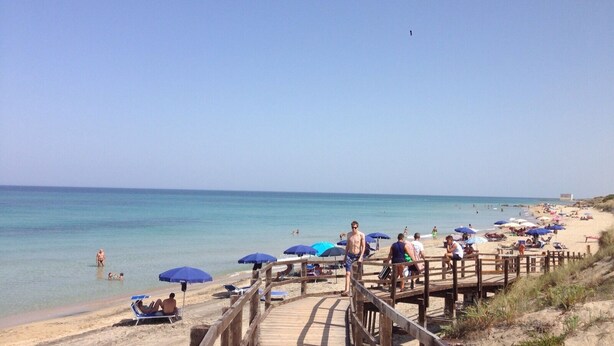 Una pasarela de madera que conduce a una playa arenosa con sombrillas azules y gente tomando el sol.
