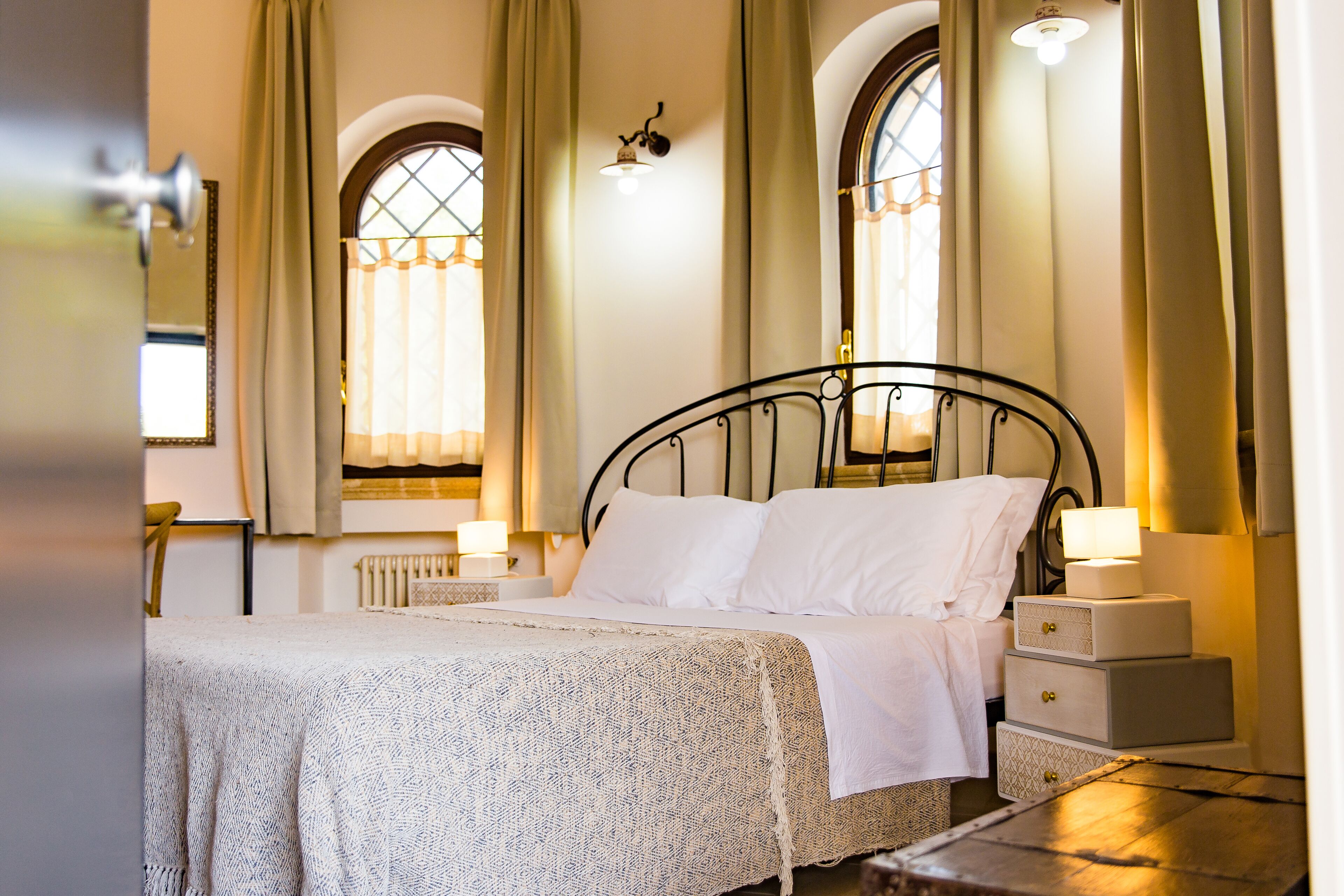 Uma cama bem arrumada com lençóis brancos, uma mesa de cabeceira com abajur e uma mesinha de cabeceira com gavetas.
