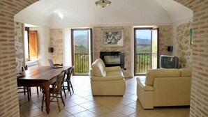 Un salon spacieux doté d’une cheminée, un coin repas avec une table et des chaises en bois, et un balcon offrant une vue sur la campagne.