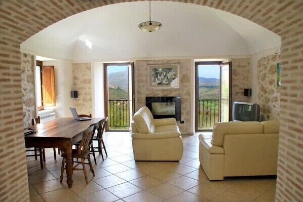 Una amplia sala de estar con chimenea, comedor con mesa y sillas de madera, y balcón con vistas al campo.
