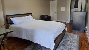 A bedroom with a bed, a desk, a chair, a rug, a door, and a small kitchenette area. - The Guest House (Circleville)