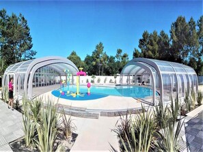 Una piscina techada con zona de juegos acuáticos, rodeada de palmeras y un cielo despejado.