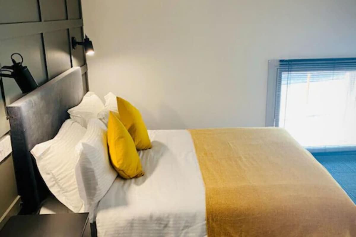 A hotel room with a bed, white and yellow pillows, a brown throw blanket, and a window with blinds.