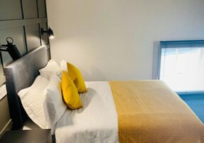 Habitación de hotel con cama, almohadas blancas y amarillas, manta marrón y ventana con persianas.