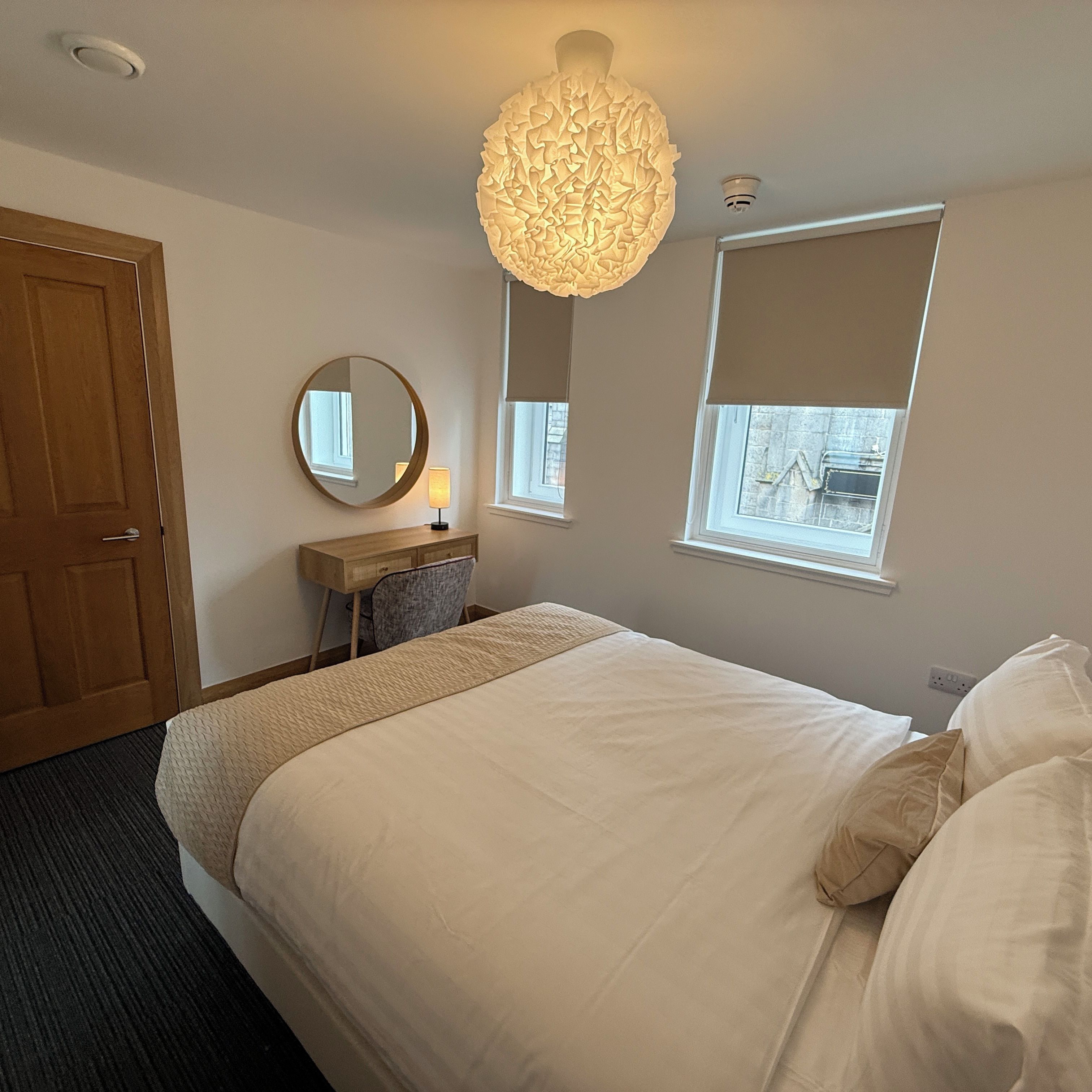 Een slaapkamer met een bed, nachtkastje, lamp, spiegel en raam.