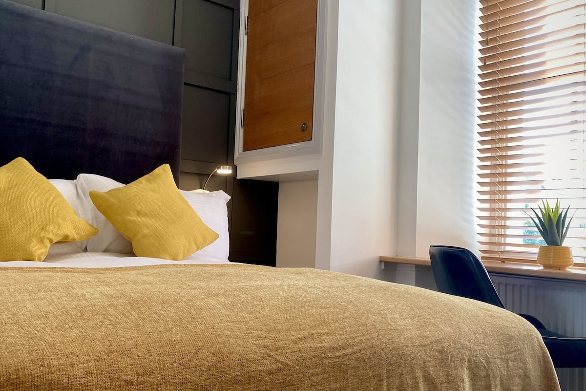 A hotel room with a large bed, two pillows, a nightstand, a lamp, a chair, and a window with blinds.