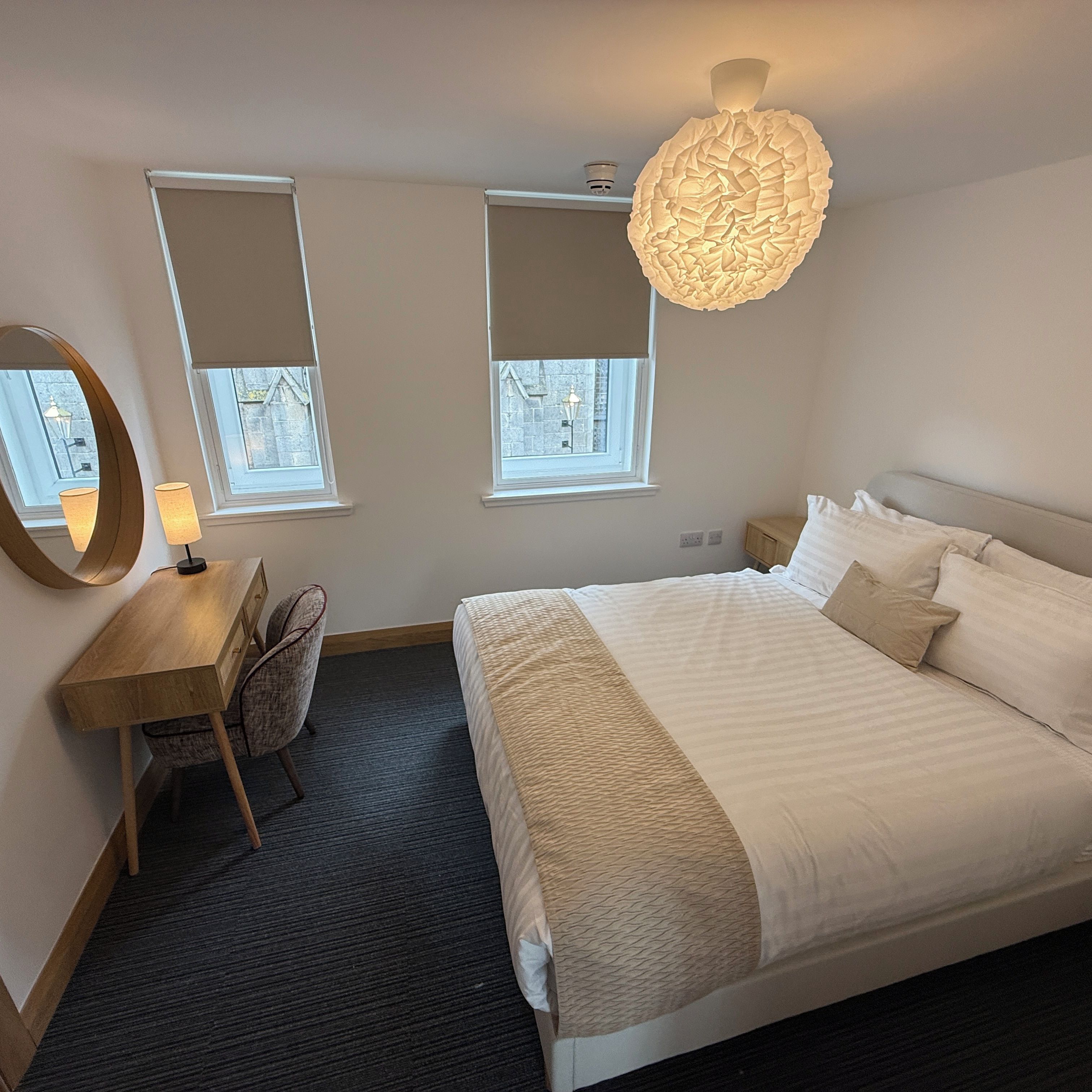 A bedroom with a bed, a desk, a chair, two windows, and a unique ceiling light fixture.