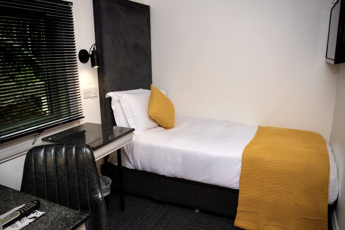 A hotel room with a bed, a desk, a chair, a window with blinds, and a wall-mounted TV.