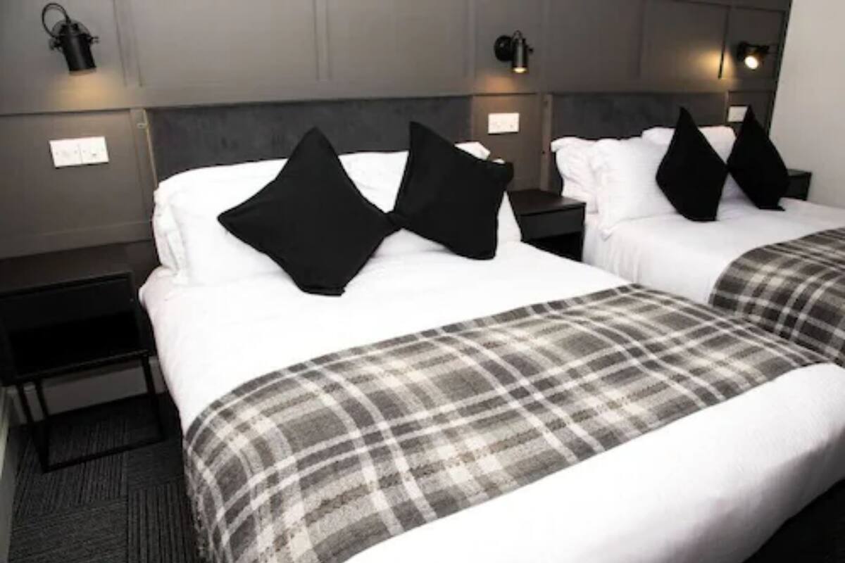 A hotel room with two beds, each with white linens and black pillows. There are two bedside tables with lamps, and a gray carpet.