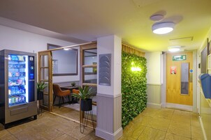 A hotel lobby with a vending machine, a seating area with chairs and a table, a green wall with plants, and a wooden door with an exit sign.