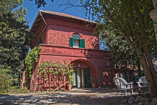 Um edifício vermelho com entrada em arco, porta verde e uma pequena janela com persianas verdes. Há plantas em vasos e uma cadeira na parte externa.