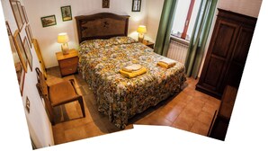 A bedroom with a floral bedspread, a wooden headboard, a nightstand with a lamp, a chair, and a window with curtains. - Holiday in a comfortable and renovated ancient villa (mottola)