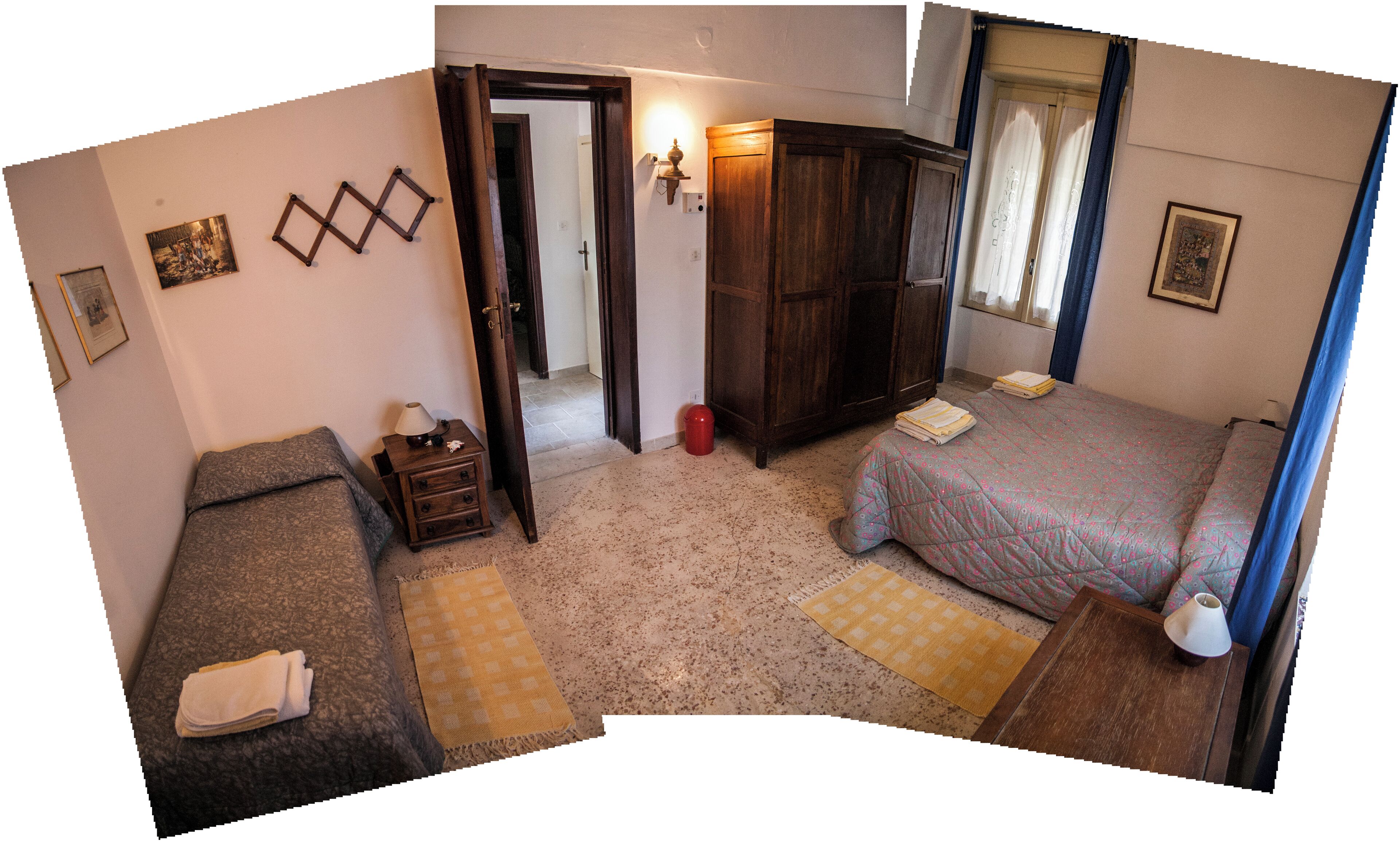 A bedroom with two single beds, a wooden wardrobe, a small bedside table with a lamp, and a door leading to another room.