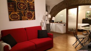 A red sofa with black cushions, a wooden floor, a kitchen area with a white cabinet, a dining table with chairs, and a large abstract painting on the wall. - L' Arcata a Montesanto 1700s courtyard, metro & funicular (Napoli)