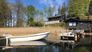 Un lago tranquilo con un bote encallado en un muelle de madera. Al fondo, se aprecia una cabaña moderna con un ventanal grande y una estructura más pequeña con un muelle.