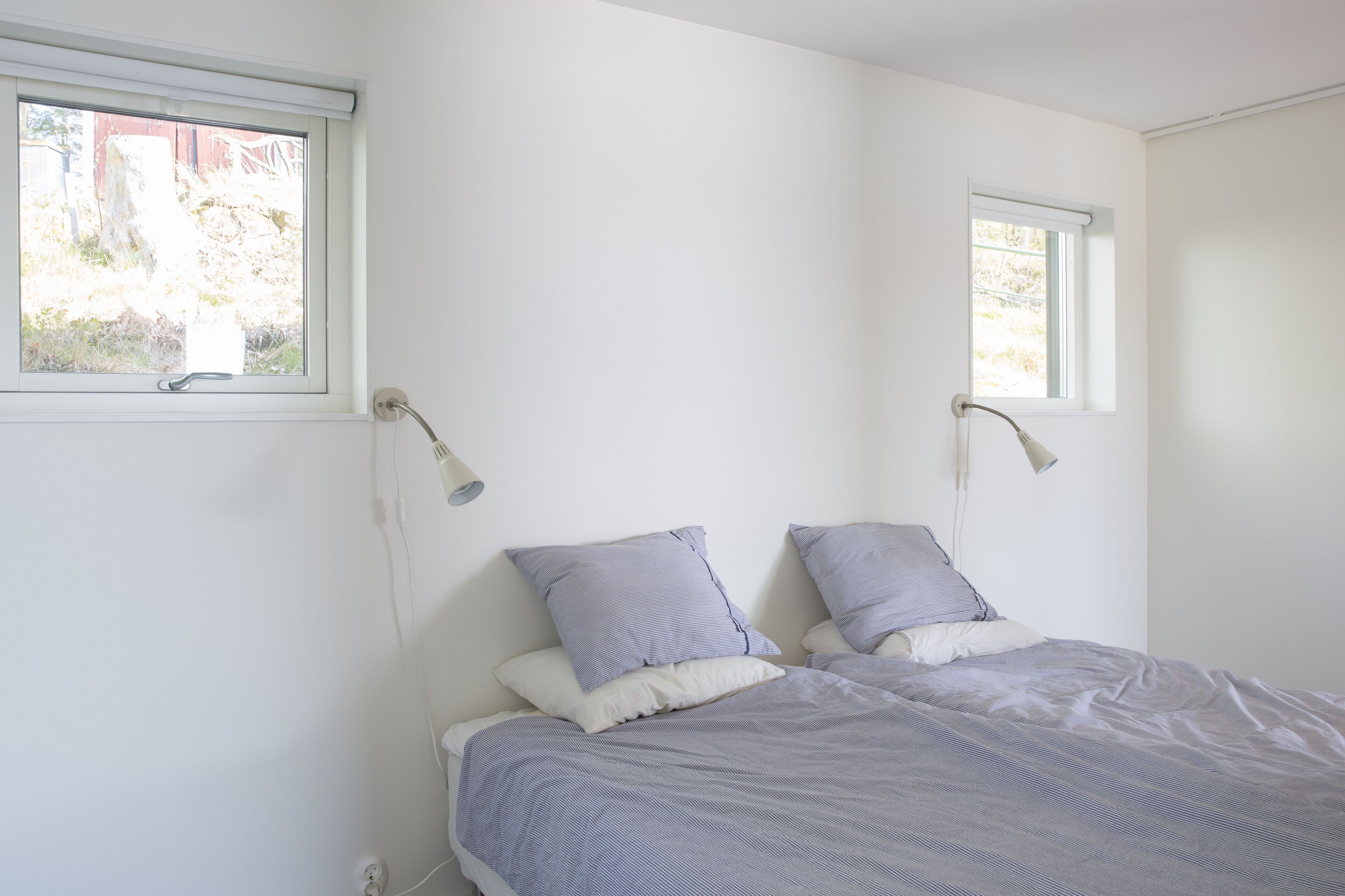 A bedroom with two beds, each with a pillow and a lamp. There are windows on the wall.